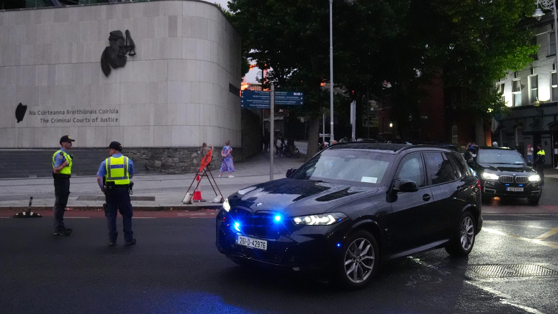 Zwei dunkle Autos einer Fahrzeugkolonne fahren auf einer Straße um die Ecke. Am Rand stehen zwei Polizisten in gelben Warnwesten. Zwei dunkle Autos einer Fahrzeugkolonne fahren auf einer Straße um die Ecke. Am Rand stehen zwei Polizisten in gelben Warnwesten.
