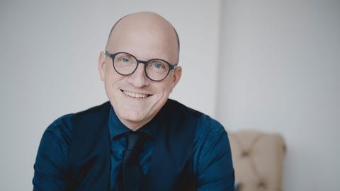 Ein Mann mit Brille und blauen Augen blickt lächelnd in die Kamera. Er trägt ein dunkelblaues Hemd, Weste und Krawatte.