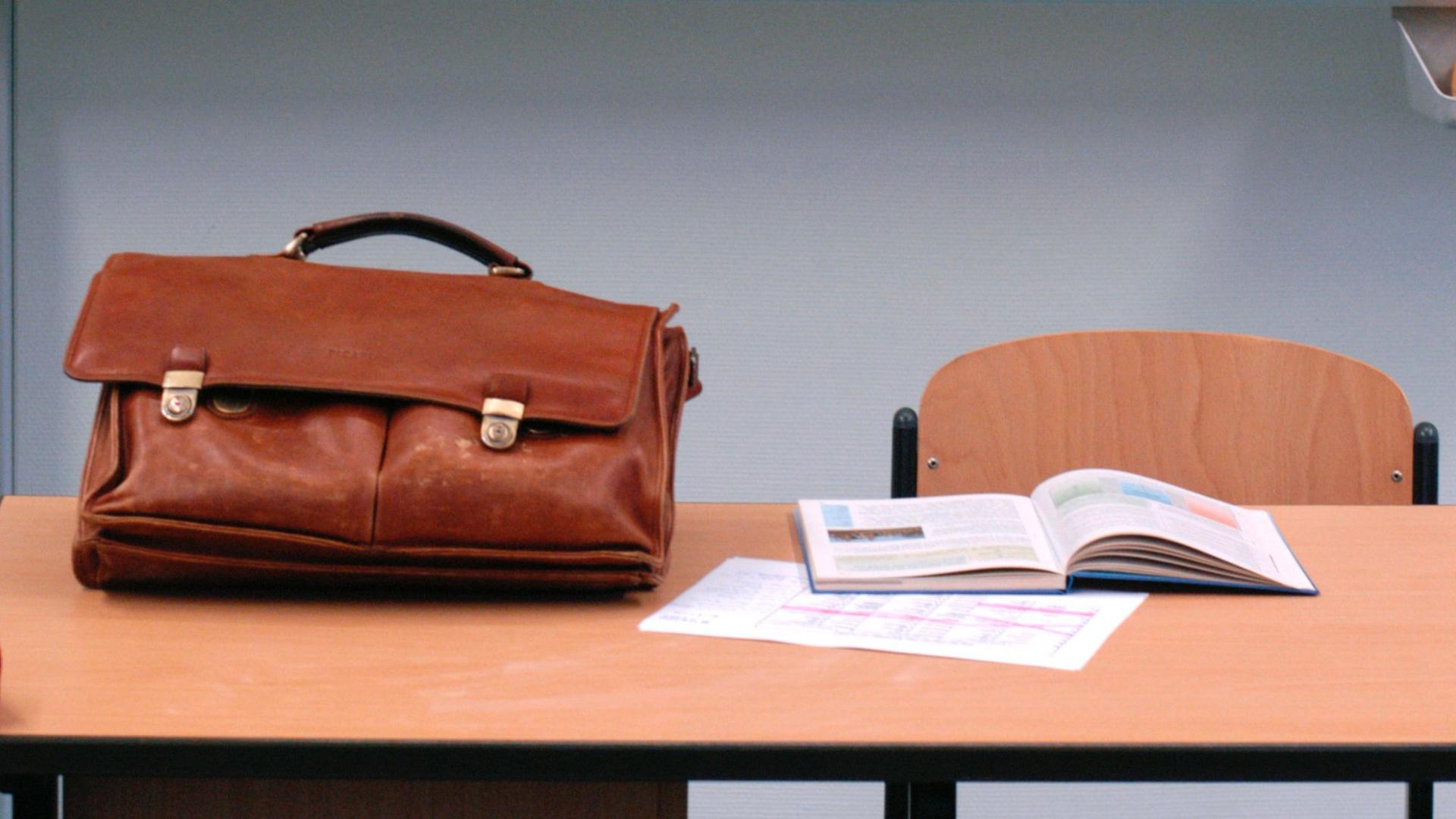 Eine Ledertasche und ein aufgeschlagenes Lehrbuch liegen auf einem leh Lehrerpult.