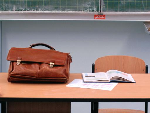Eine Ledertasche und ein aufgeschlagenes Lehrbuch liegen auf einem Lehrerpult.