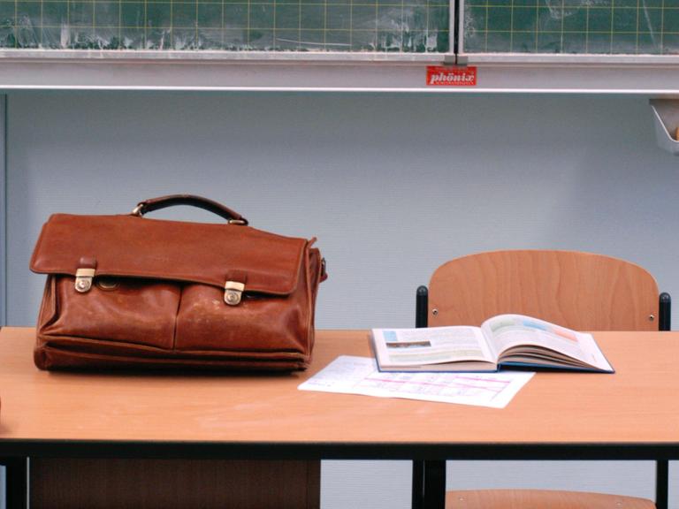 Eine Ledertasche und ein aufgeschlagenes Lehrbuch liegen auf einem Lehrerpult.
