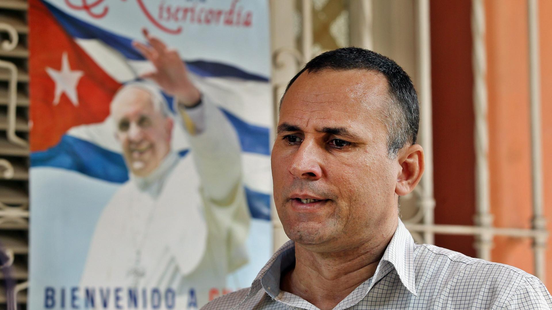 Ferrer steht vor einem vergitterten Fenster, an dem ein Plakat mit dem Bild von Papst Franziskus vor der kubanischen Flagge hängt.