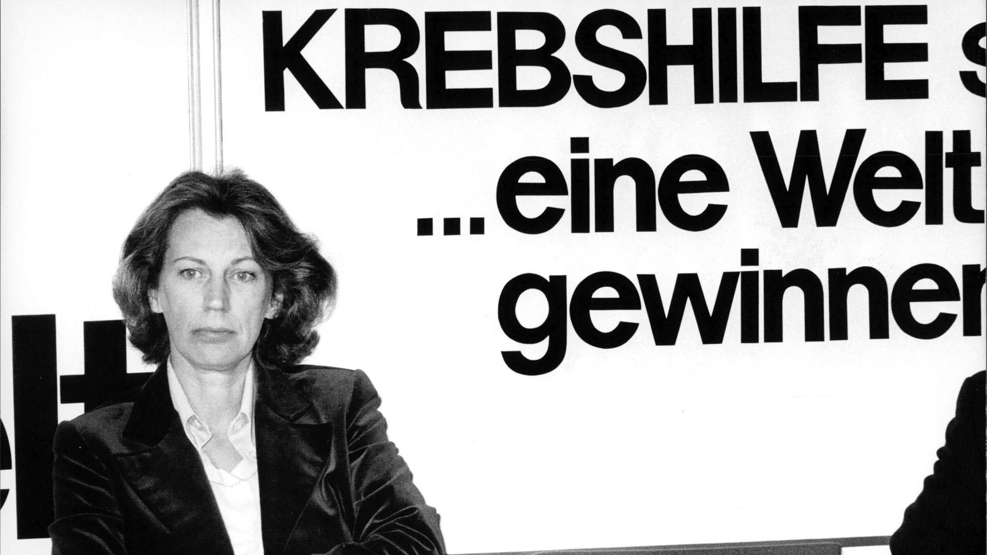 Mildred Scheel ist in einer Schwarz-weiß-Aufnahme mit verschrenkten Armen vor einer Wand mit der Aufschrift "Krebshilfe" zu sehen Mildred Scheel ist in einer Schwarz-weiß-Aufnahme mit verschrenkten Armen vor einer Wand mit der Aufschrift "Krebshilfe" zu sehen