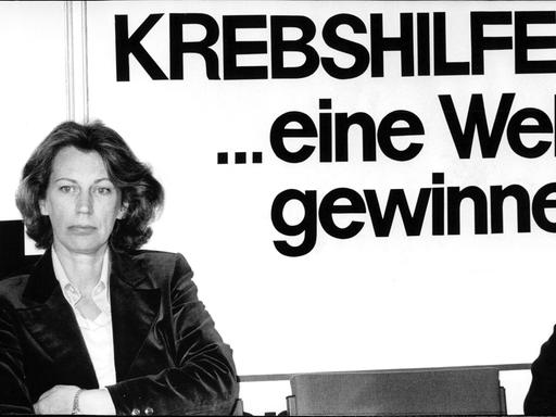 Mildred Scheel ist in einer Schwarz-weiß-Aufnahme mit verschrenkten Armen vor einer Wand mit der Aufschrift "Krebshilfe" zu sehen