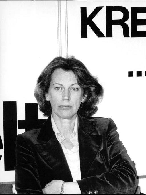 Mildred Scheel ist in einer Schwarz-weiß-Aufnahme mit verschrenkten Armen vor einer Wand mit der Aufschrift "Krebshilfe" zu sehen