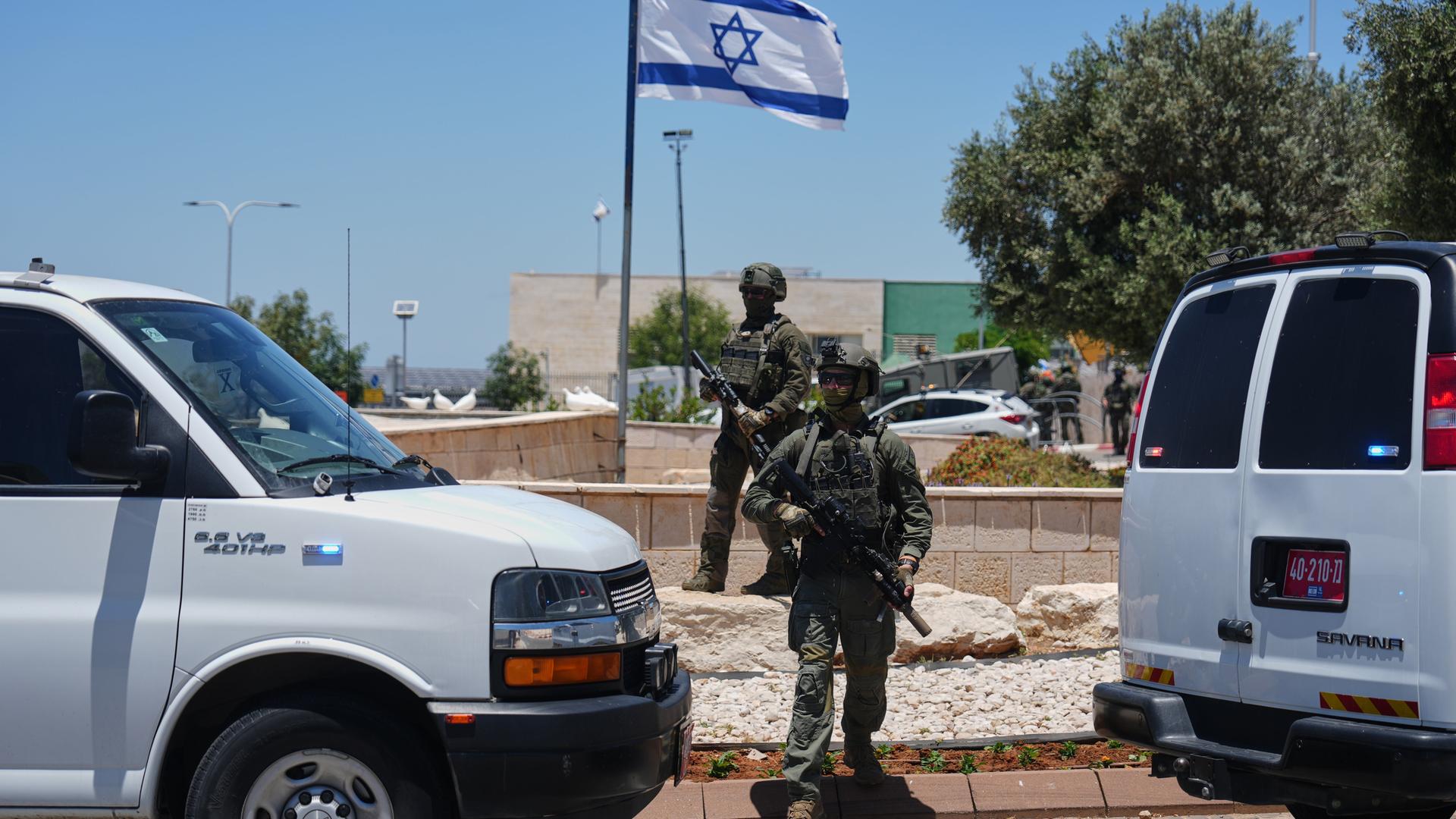Zwei weiße Fahrzeuge stehen an einer Verkehrsinsel mit einem Fahnenmast mit israelischer Flagge. Darauf stehen zwei bewaffnete Soldaten. 