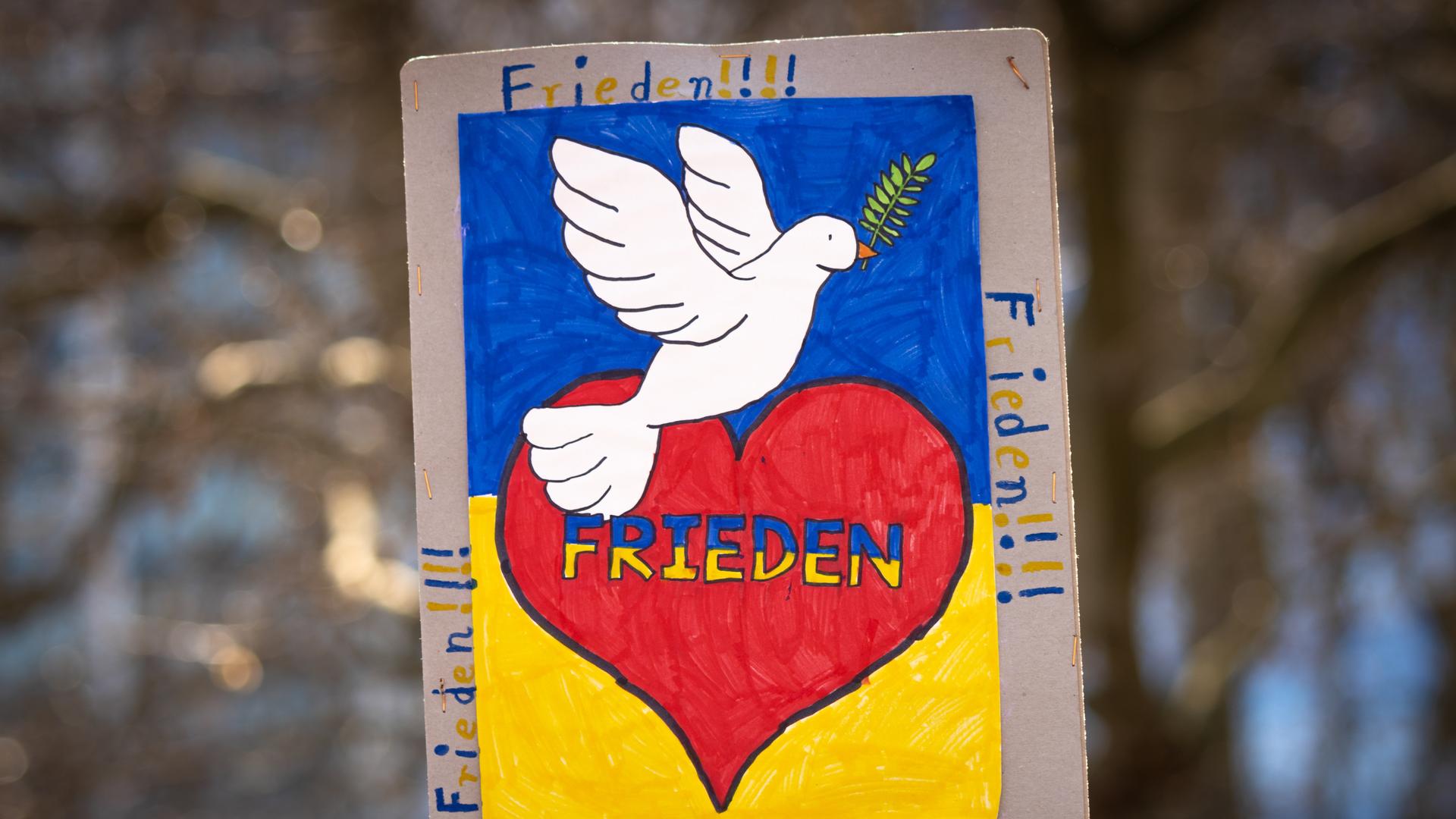 "FRIEDEN" steht auf einem Plakat. Außerdem ist ein rotes Herz und eine weiße Taube darauf zu sehen.