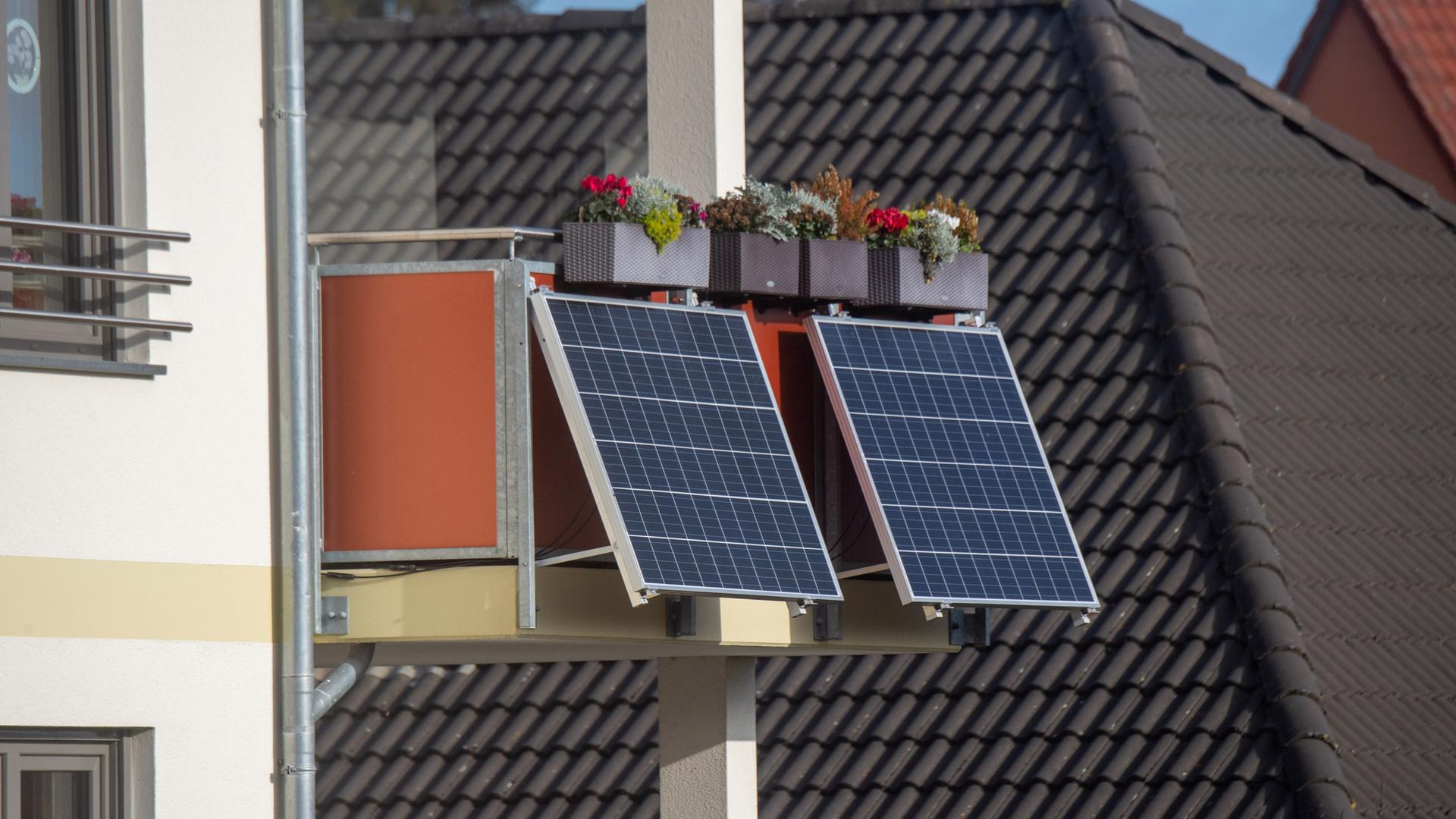 Solarmodule für ein sogenanntes Balkonkraftwerk hängen an einem Balkon unter bunt bepflanzten Blumenkästen.
