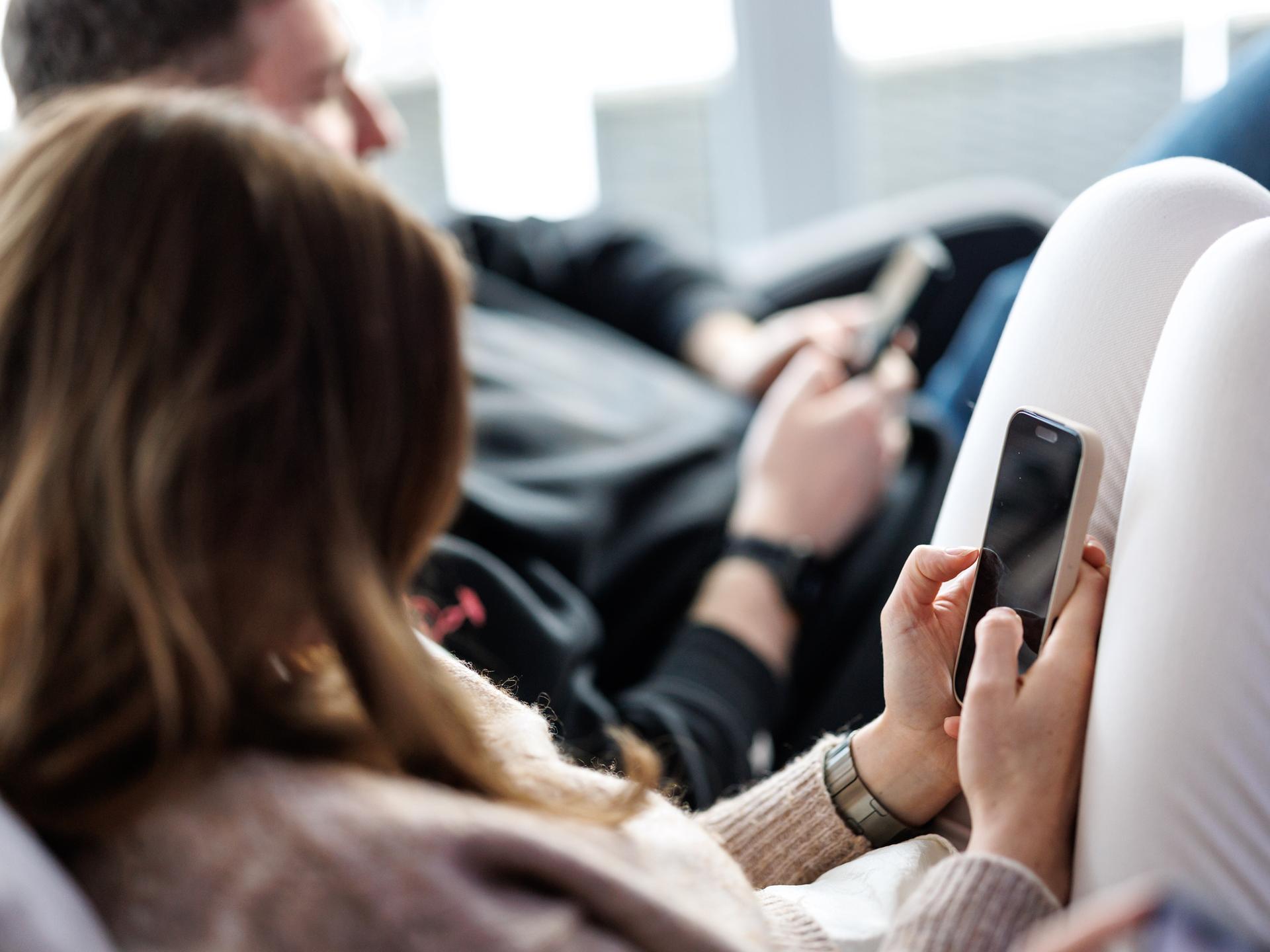Eine junge Frau sitzt auf einem Sofa und benutzt ein Smartphone. Neben ihr sitzt ein junger Mann, der ebenfalls am Handy tippt. 