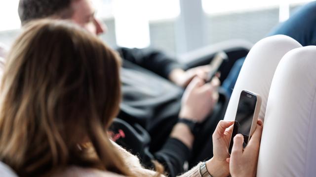 Eine junge Frau sitzt auf einem Sofa und benutzt ein Smartphone. Neben ihr sitzt ein junger Mann, der ebenfalls am Handy tippt. 
