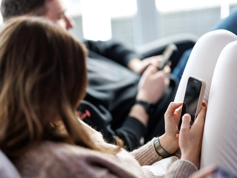Eine junge Frau sitzt auf einem Sofa und benutzt ein Smartphone. Neben ihr sitzt ein junger Mann, der ebenfalls am Handy tippt. 