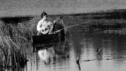 Schwarzweißbild von 1976: Ein Mitglied des Praslovo Posidelki (Praslovo Gatherings) Gesangsensembles aus der Region Moskau sitzt am Wasser und spielt Gitarre.