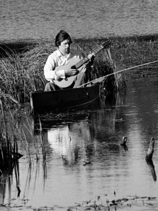 Schwarzweißbild von 1976: Ein Mitglied des Praslovo Posidelki (Praslovo Gatherings) Gesangsensembles aus der Region Moskau sitzt am Wasser und spielt Gitarre.
