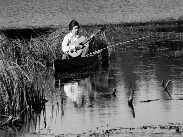 Schwarzweißbild von 1976: Ein Mitglied des Praslovo Posidelki (Praslovo Gatherings) Gesangsensembles aus der Region Moskau sitzt am Wasser und spielt Gitarre.