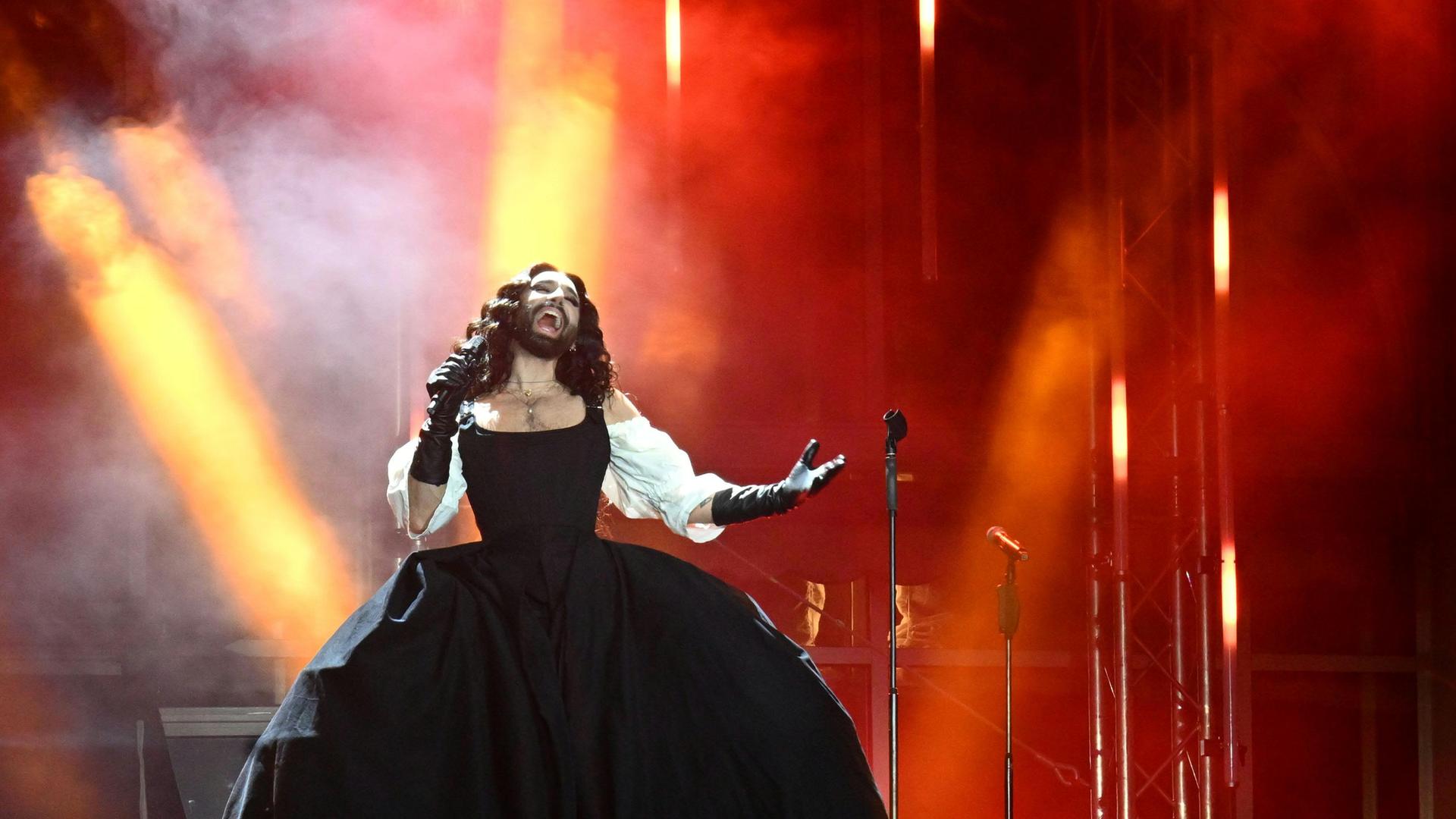Conchita Wurst steht im langen Kleid auf einer rot und gelb beleuchteten Bühne und singt. Conchita Wurst steht im langen Kleid auf einer rot und gelb beleuchteten Bühne und singt.