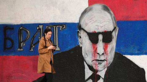 Eine Frau geht an einem Wandbild von Putin vorbei, das mit einer schwarzen Sonnenbrille und daraus tropfendem Blut übermalt wurde. Daneben steht das Wort "Bruder".