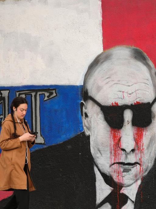 Eine Frau geht an einem Wandbild von Putin vorbei, das mit einer schwarzen Sonnenbrille und daraus tropfendem Blut übermalt wurde. Daneben steht das Wort "Bruder".