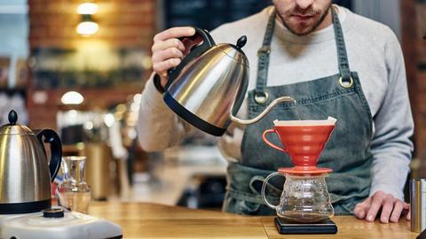 Ein Barista mit Schürze gießt kochendes Wasser in einen Kaffeefilter in einem Coffeeshop.