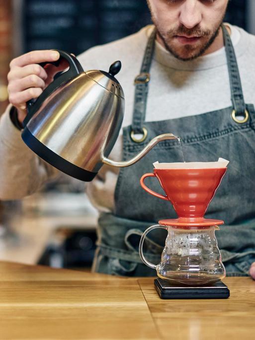Ein Barista mit Schürze gießt kochendes Wasser in einen Kaffeefilter in einem Coffeeshop.