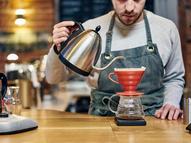 Ein Barista mit Schürze gießt kochendes Wasser in einen Kaffeefilter in einem Coffeeshop.