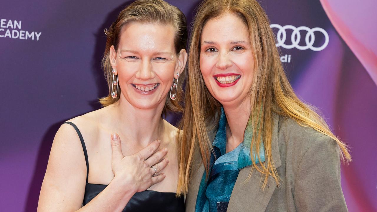 Schauspielerin Sandra Hüller und Regisseurin Justine Triet stehen elegant angezogen nah nebeneinander und lachen in die Kamera. Hüller hält eine Hand an ihr Dekolletee.