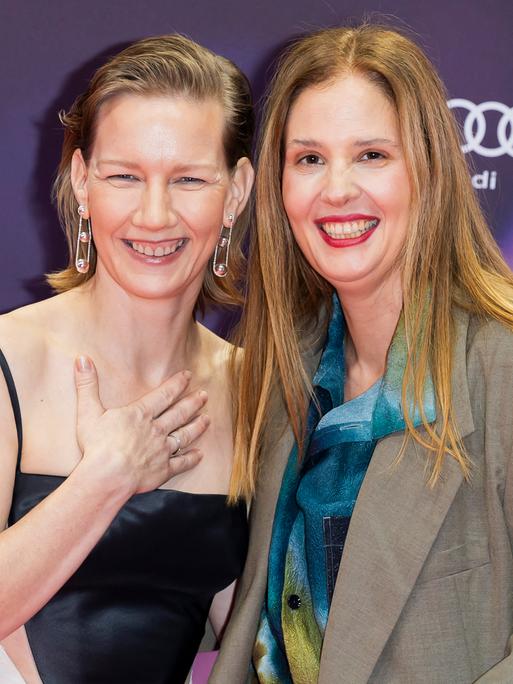 Schauspielerin Sandra Hüller und Regisseurin Justine Triet stehen elegant angezogen nah nebeneinander und lachen in die Kamera. Hüller hält eine Hand an ihr Dekolletee.