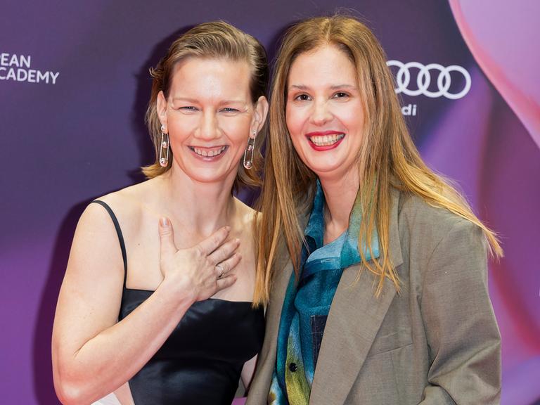 Schauspielerin Sandra Hüller und Regisseurin Justine Triet stehen elegant angezogen nah nebeneinander und lachen in die Kamera. Hüller hält eine Hand an ihr Dekolletee.