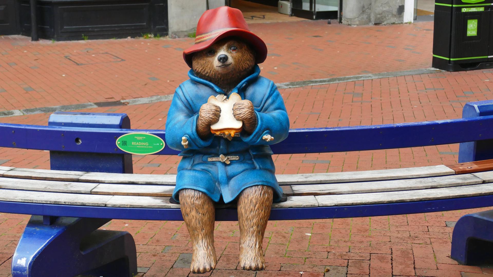Eine Statue der Kinderbuchfigur Paddington Bär sitzt auf einer Bank. Sie hält ein Sandwich in der Hand. Eine Statue der Kinderbuchfigur Paddington Bär sitzt auf einer Bank. Sie hält ein Sandwich in der Hand.