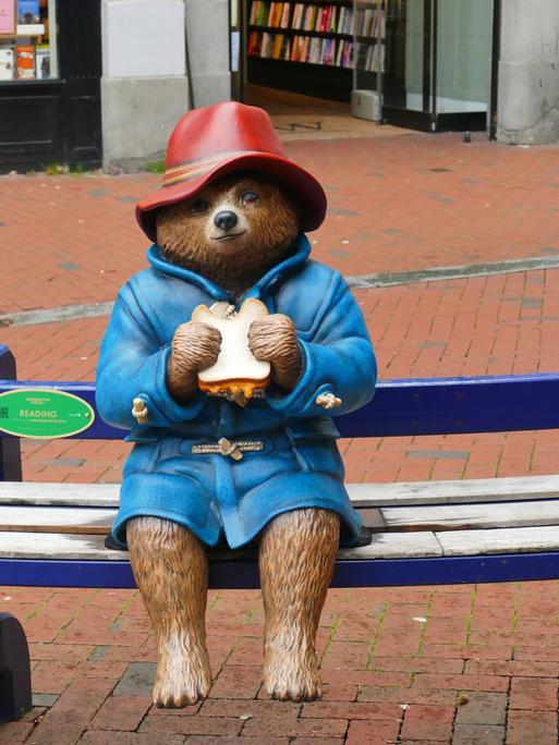 Eine Statue der Kinderbuchfigur Paddington Bär sitzt auf einer Bank. Sie hält ein Sandwich in der Hand.