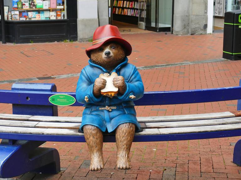 Eine Statue der Kinderbuchfigur Paddington Bär sitzt auf einer Bank. Sie hält ein Sandwich in der Hand.