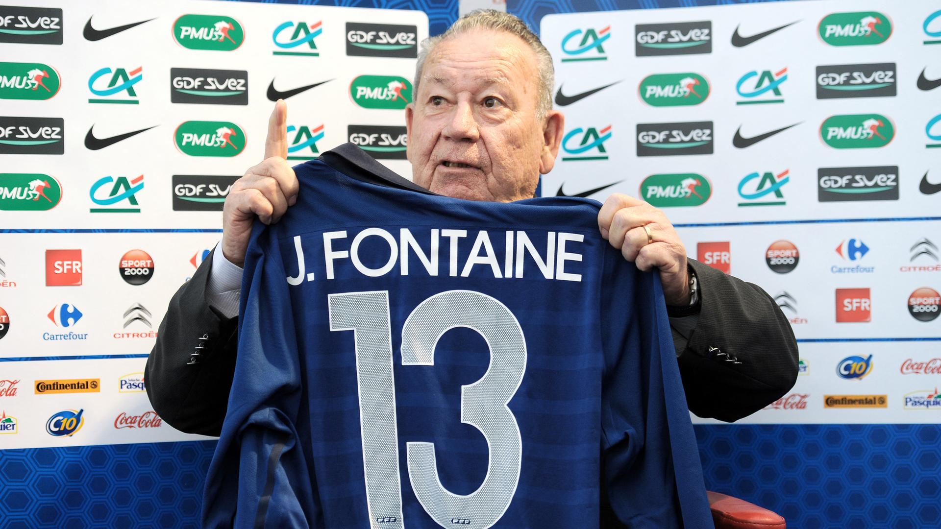 Das Foto zeigt den ehemaligen Fußballspieler Just Fontaine, der ein blaues Trikot mit dem Namen Fontaine und der Nummer 13 hochhält.
