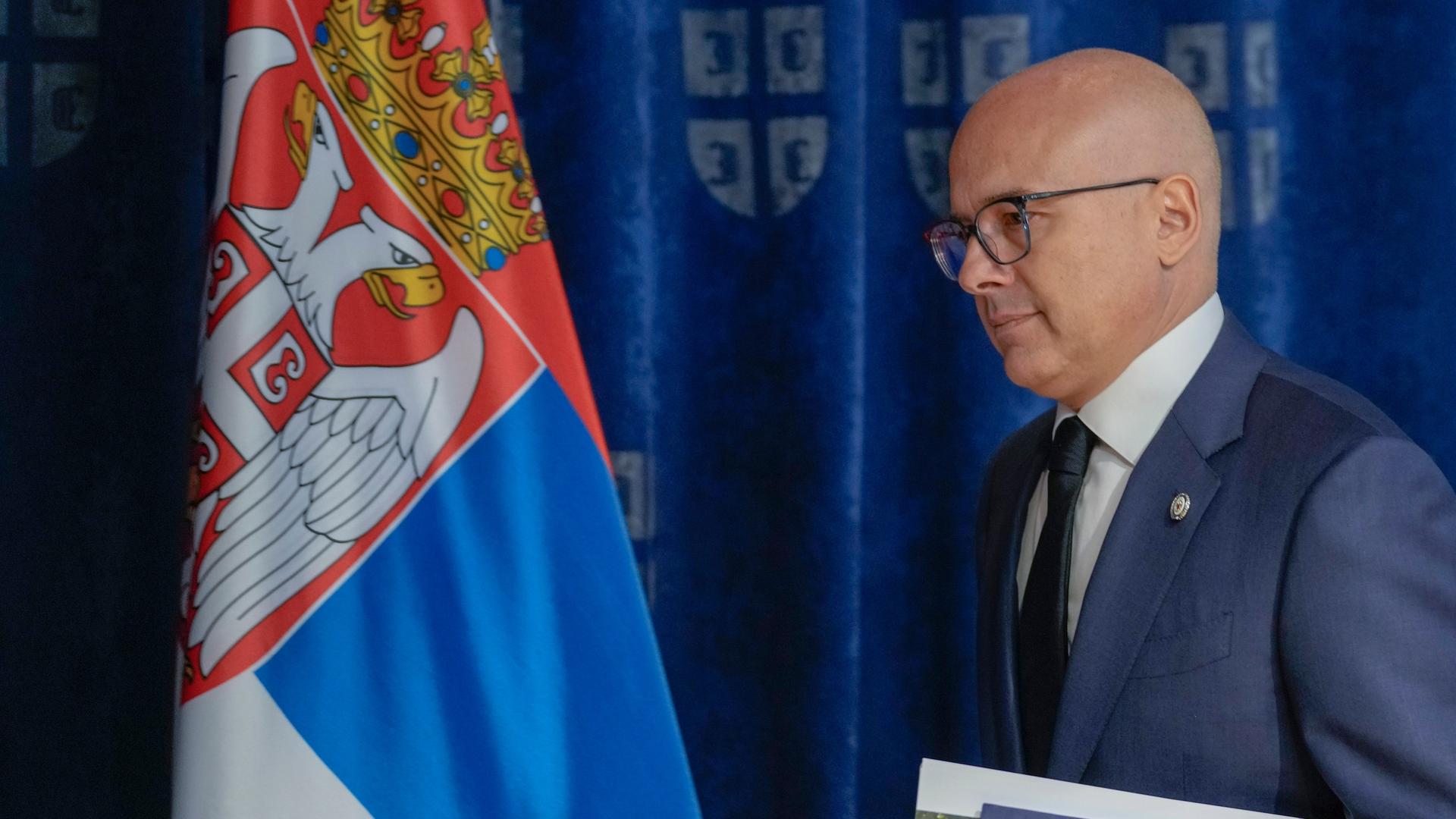 Serbiens Ministerpräsident Milos Vucevic ist von der Seite fotografiert. Er hält Unterlagen in der Hand. Im Hintergrund ist die serbische Flagge. 