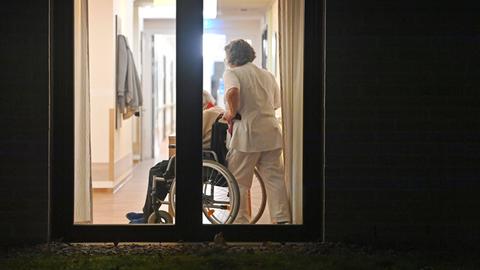 Blick in ein Seniorenheim durch ein Fenster: Eine Pflegekraft schiebt jemanden in einem Rollstuhl.