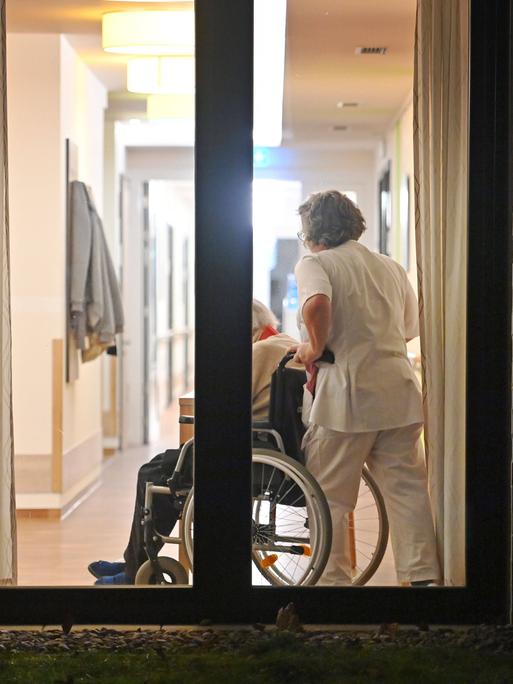 Blick in ein Seniorenheim durch ein Fenster: Eine Pflegekraft schiebt jemanden in einem Rollstuhl.