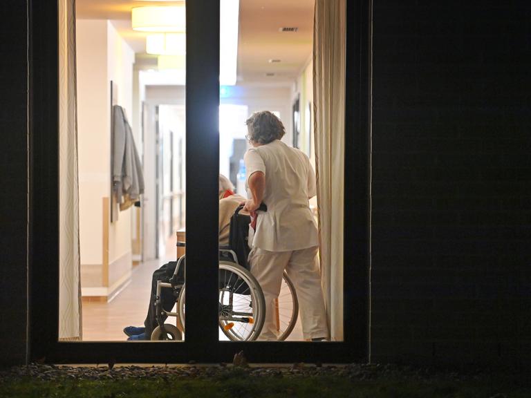 Blick in ein Seniorenheim durch ein Fenster: Eine Pflegekraft schiebt jemanden in einem Rollstuhl.