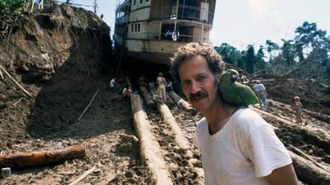 Werner Herzog am Set zu "Fitzcarraldo" mit einem grünen Papageien auf der Schulter und einem auf Baumstämmen transportierten Dampfschiff im Hintergrund.