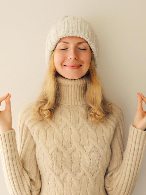 Lächelnde junge Frau meditiert in Yogaposition in winterlich warmem Strickpullover und Mütze