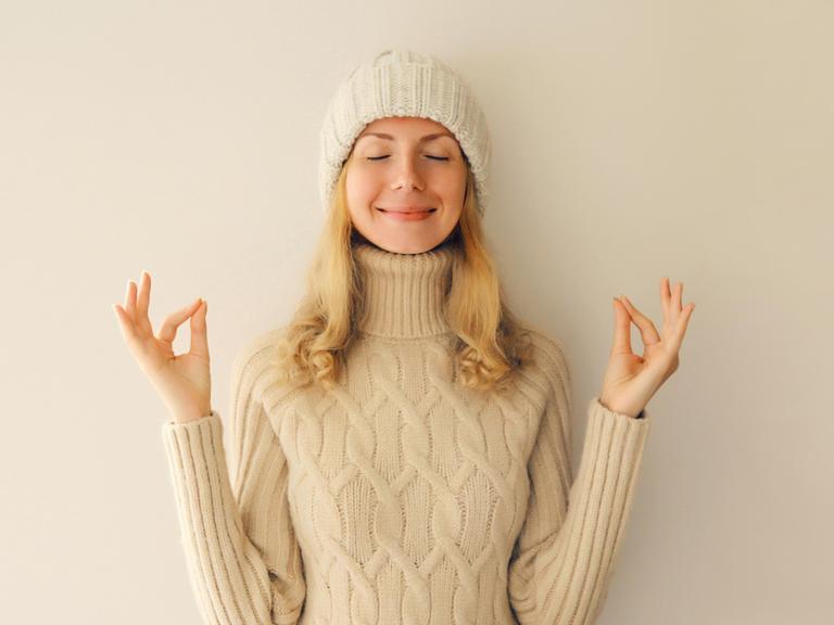 Lächelnde junge Frau meditiert in Yogaposition in winterlich warmem Strickpullover und Mütze