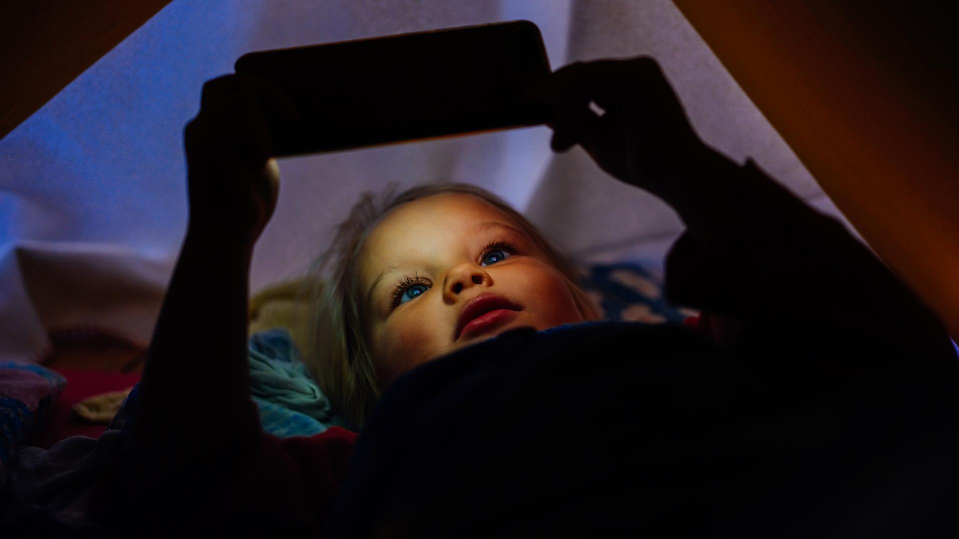 Ein Kleines Mädchen, das mit ihrem Handy im Bett etwas ansieht. Der Lichtschein des Bildschirms beleuchtet ihr Gesicht.