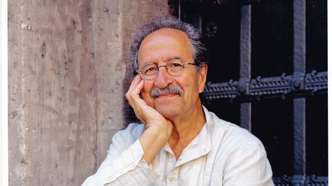 Ein Mann mit kurzen grauen Haaren und Schnurrbart sitzt in einer Fensternische. Er lächelt freundlich und hat eine runde Brille. Das eine Bein ist angezogen, den Kopf stützt er in die Hand. Er trägt ein weißes Leinenhemd und -hose. 