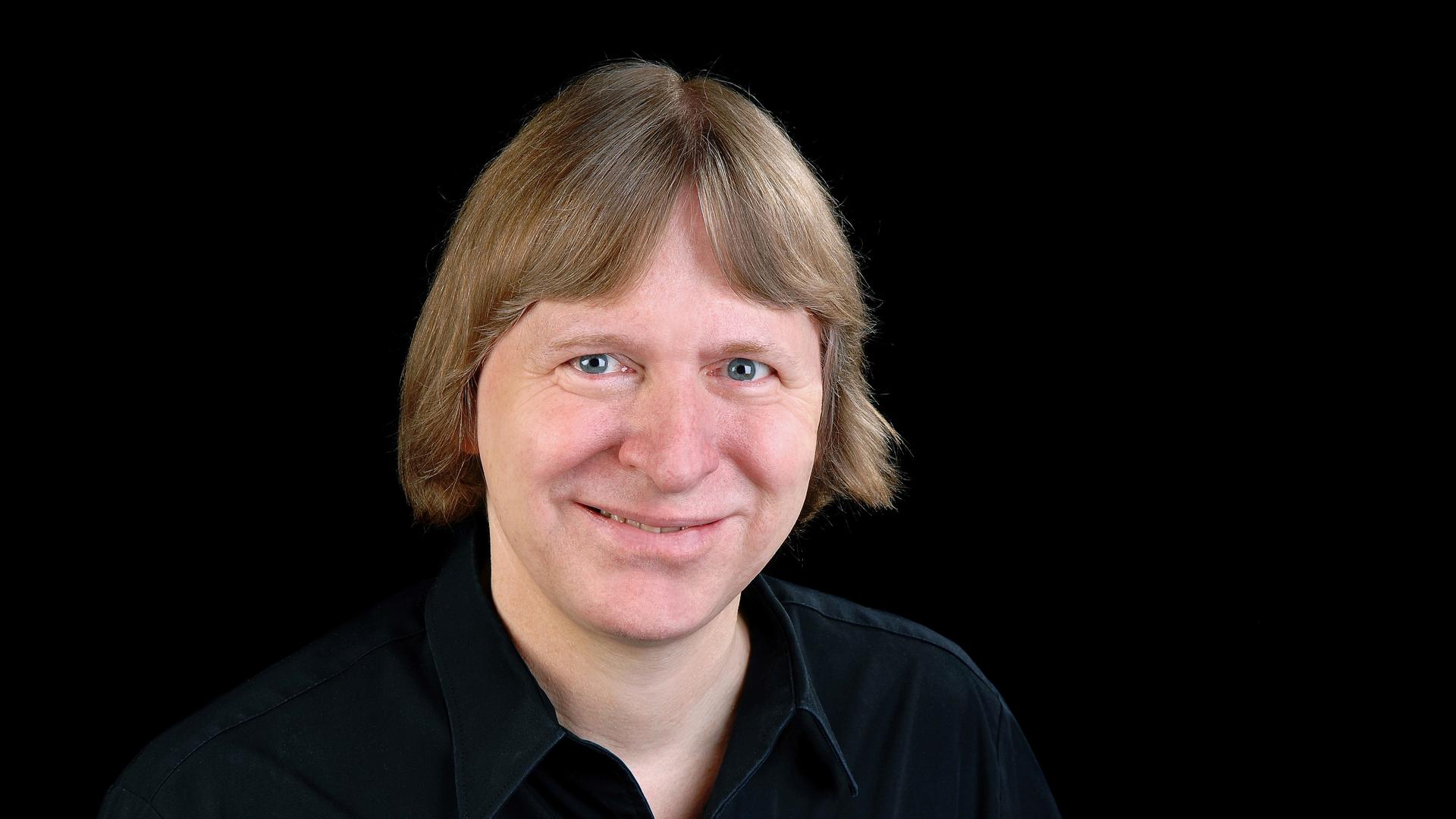 Ein Mann mit halblangen Haaren vor dunklem Hintergrund schaut lächelnd in die Kamera