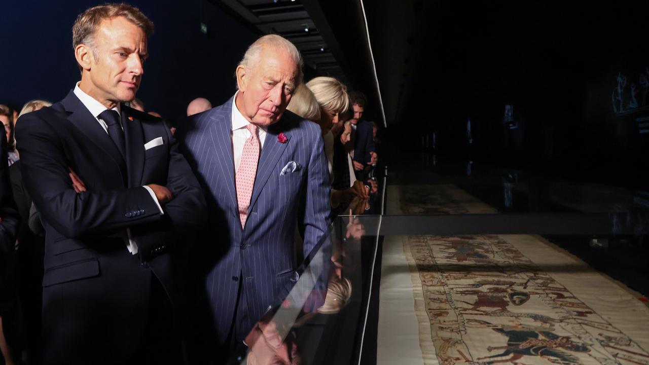 Der französische Präsident Macron und König Charles schauen sich den unter Glas liegenden Teppich an.