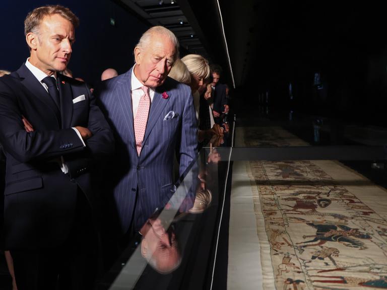 Der französische Präsident Macron und König Charles schauen sich den unter Glas liegenden Teppich an.