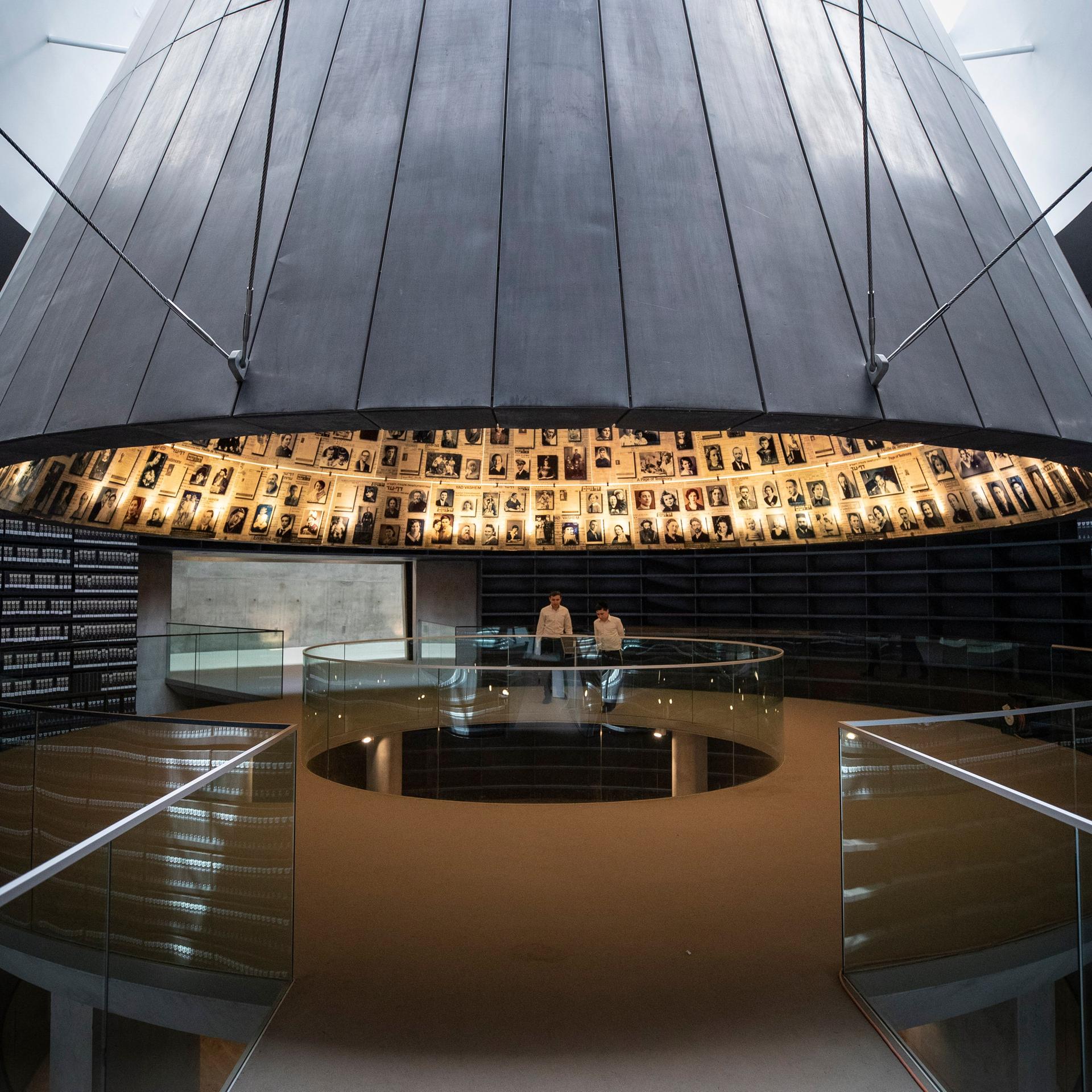 Besucher in einer dunklen Halle unter einer fast raumfüllenden Stahlglocke, deren Innenseite mit Schwarz-Weiß-Porträts ausgekleidet ist. 