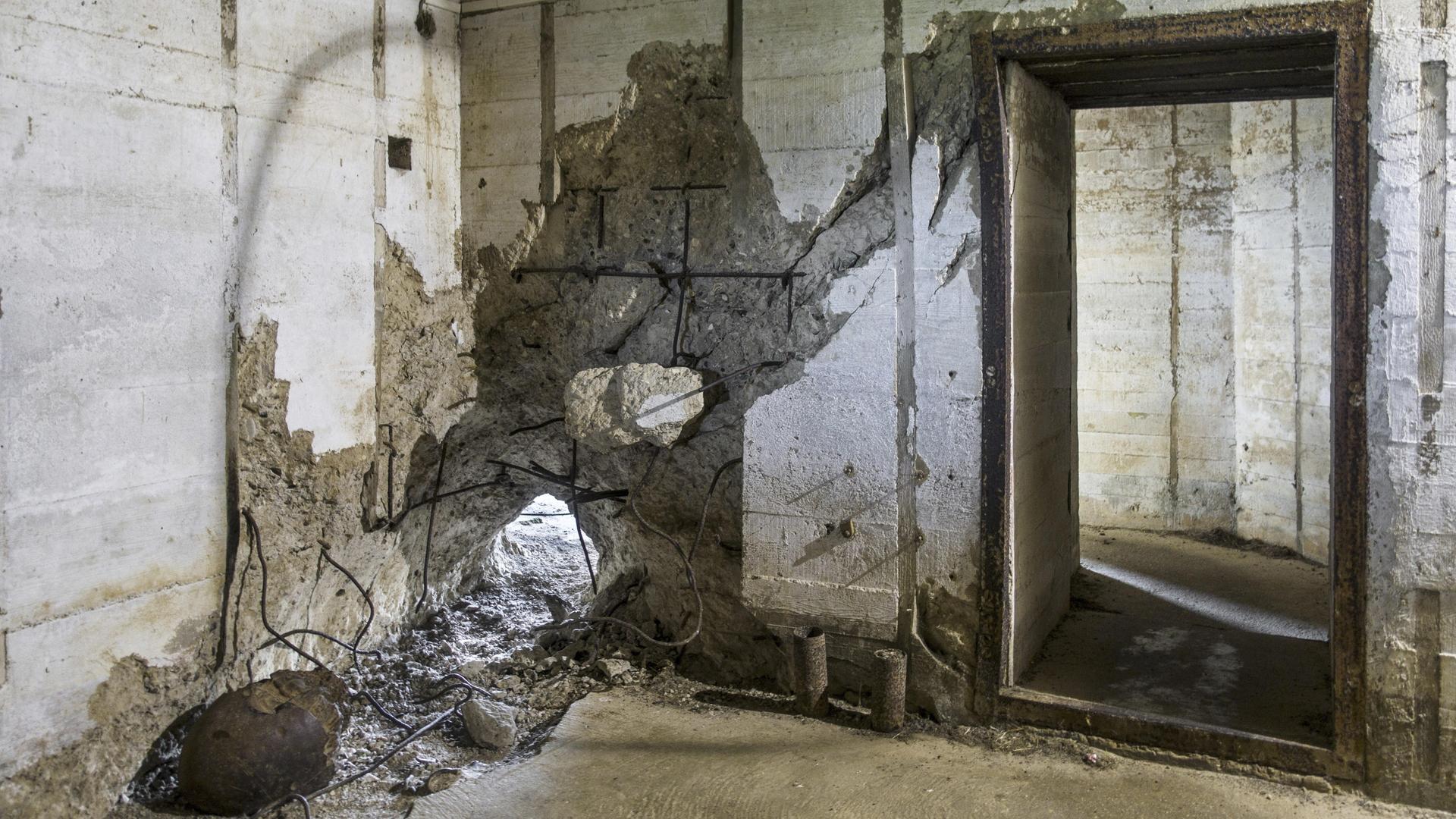 Raum in einem Bunker, in dem eine Betonwand an einer Stelle durchschlagen ist.