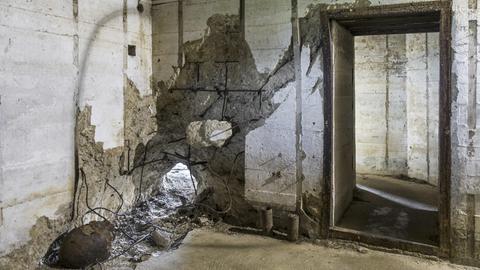 Raum in einem Bunker, in dem eine Betonwand an einer Stelle durchschlagen ist.