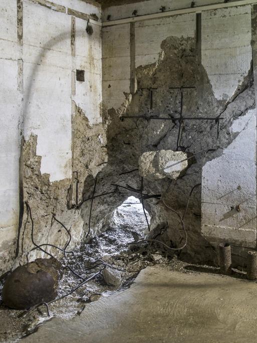 Raum in einem Bunker, in dem eine Betonwand an einer Stelle durchschlagen ist.