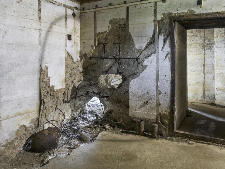 Raum in einem Bunker, in dem eine Betonwand an einer Stelle durchschlagen ist.