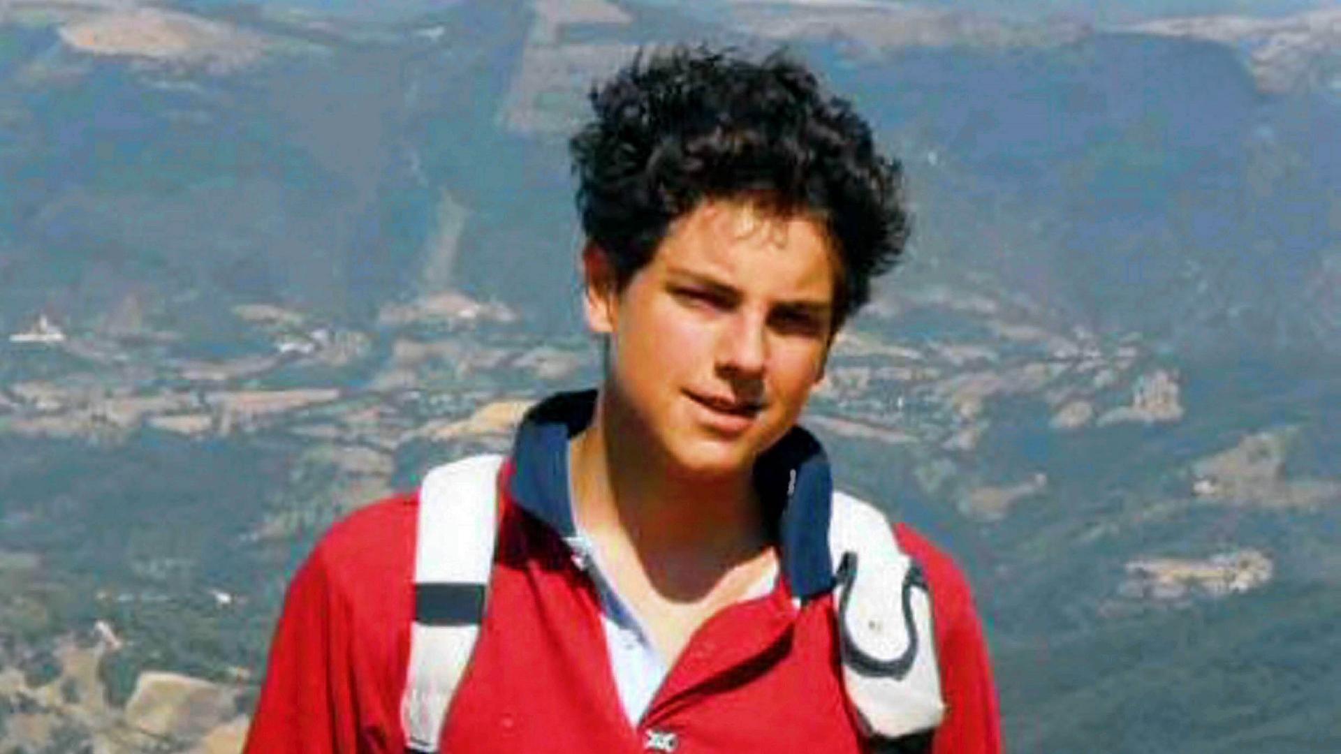 Carlo Acutis, ein Jugendlicher mit Rucksack und dunklen Haaren, steht auf einem Gebirge. 