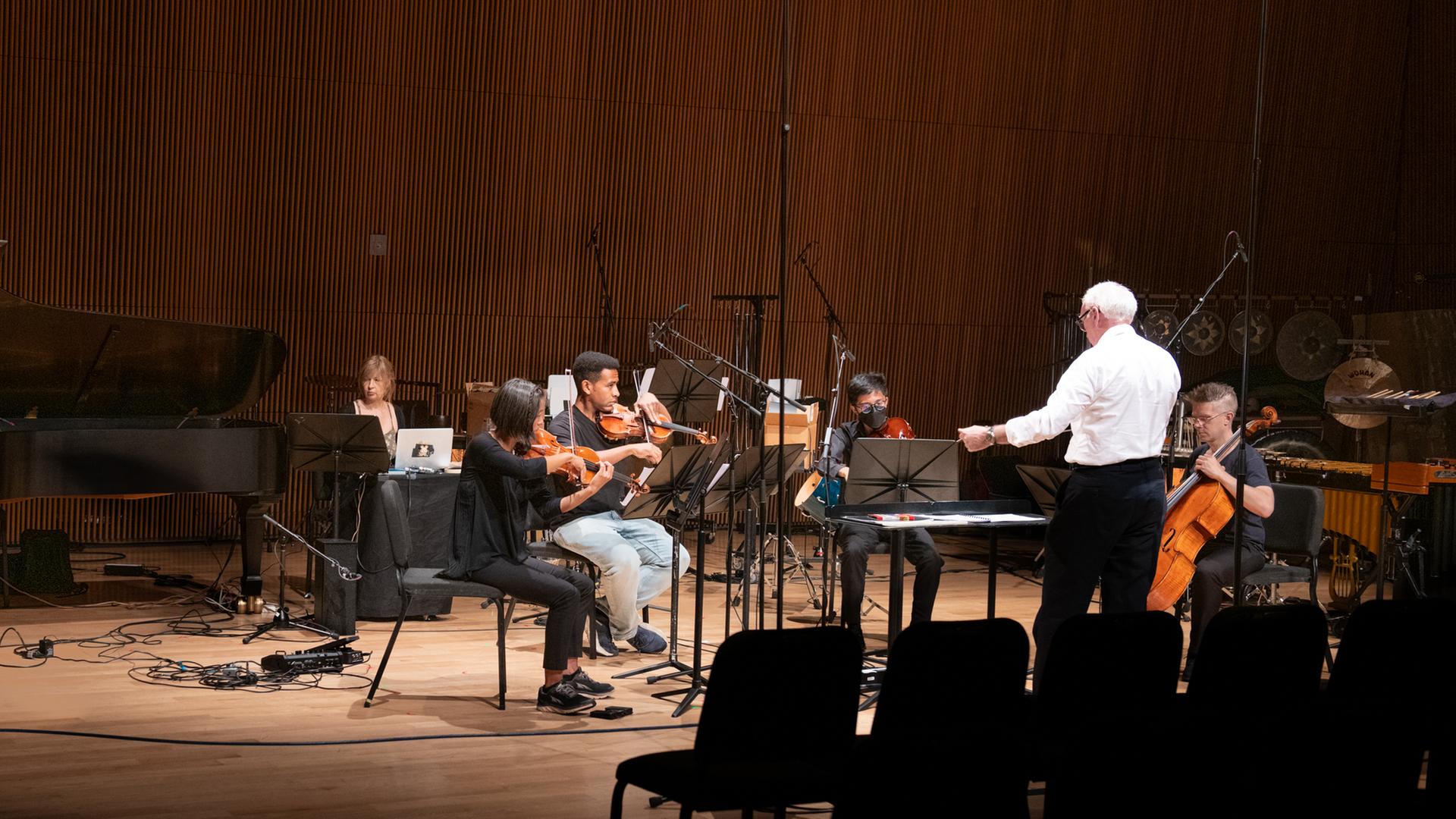 Ein Mann in weißem Hemd dirigiert ein Ensemble von 5 Musikern auf einer Bühne mit braunem Parkettboden